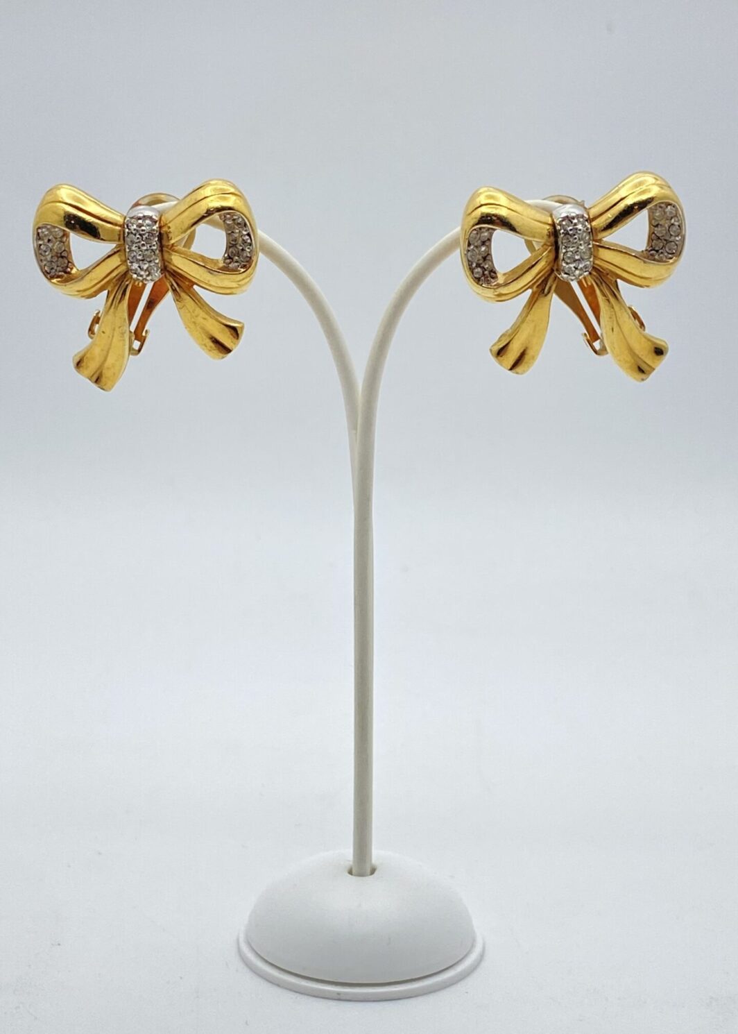 “BUTLER & WILSON”. ORECCHINI A FORMA DI FIOCCHI O NODI D’AMORE, VINTAGE, IN METALLO GOLDTONE E CRISTALLI DIAMOND LIKE. U.K. ANNI ‘70.