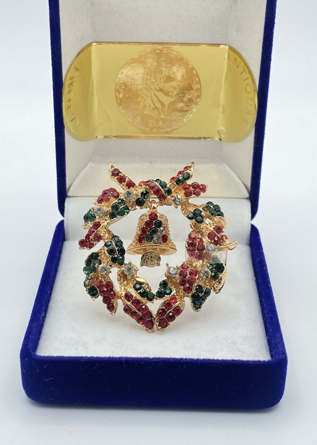 SPILLA NATALIZIA A FORMA DI GHIRLANDA, IN METALLO GOLDTONE, CRISTALLI POLICROMI E CAMPANA BASCULANTE. BIGIOTTERIA AMERICANA VINTAGE ANNI ‘80.